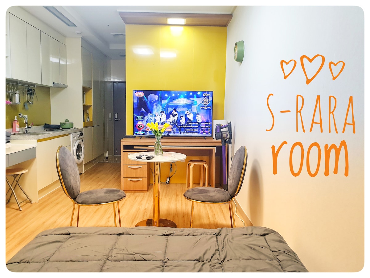 A cozy living space features a round dining table with two upholstered chairs, positioned in front of a 55-inch smart TV. Bright yellow walls and natural light contribute to the inviting atmosphere. A bed with a neutral-colored blanket is situated in the foreground.