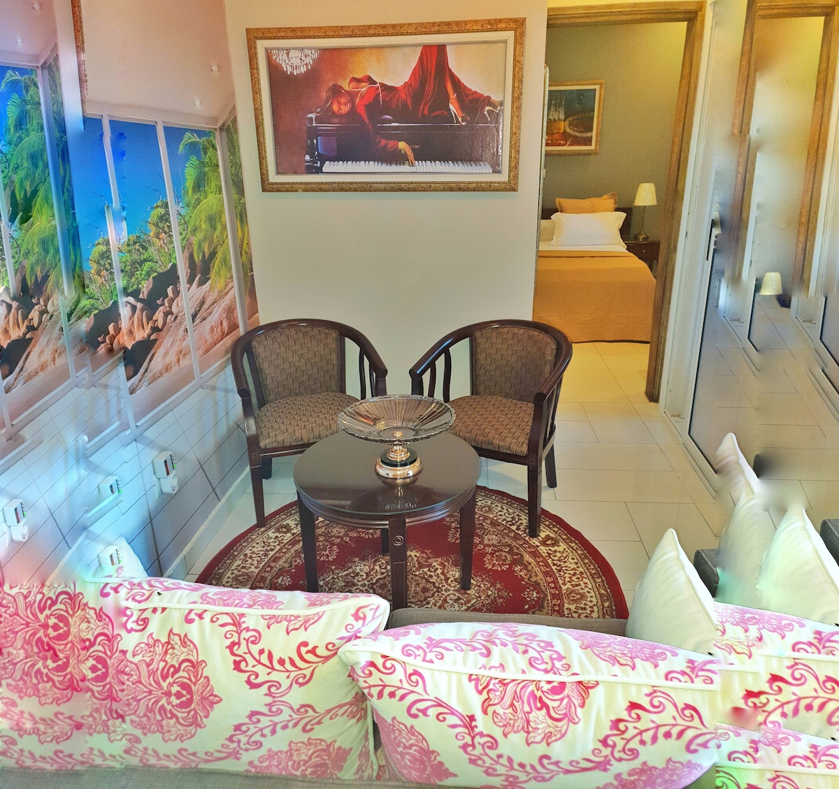 A comfortable seating area is arranged with two woven chairs and a round table, placed on a decorative rug. Behind, a bedroom is visible, featuring a spacious bed. A wall mural depicting a tropical scene adds a vibrant touch to the space.