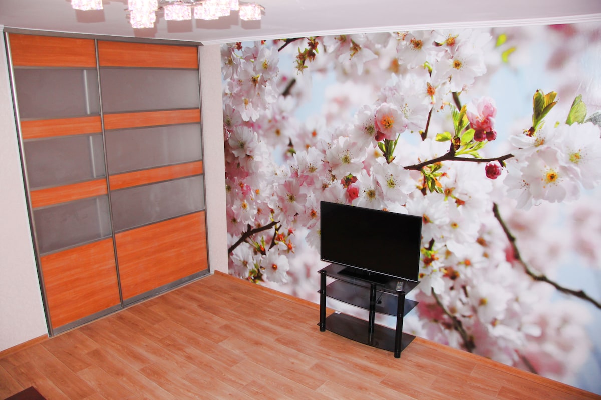 A living area features a television on a black stand, positioned in front of a wall adorned with a floral mural of cherry blossoms. Wooden storage cabinets with sliding glass doors are visible to the left, complemented by a warm wooden floor.