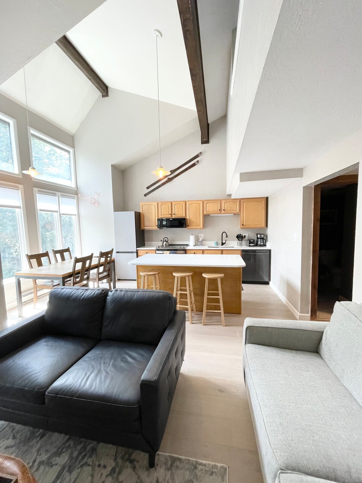 An open-concept living area is presented, showcasing high ceilings with exposed beams. A modern kitchen features wooden cabinets and a breakfast bar with stools. A dining table is visible, surrounded by chairs. Comfortable seating is provided by a dark sofa and a light-colored couch.
