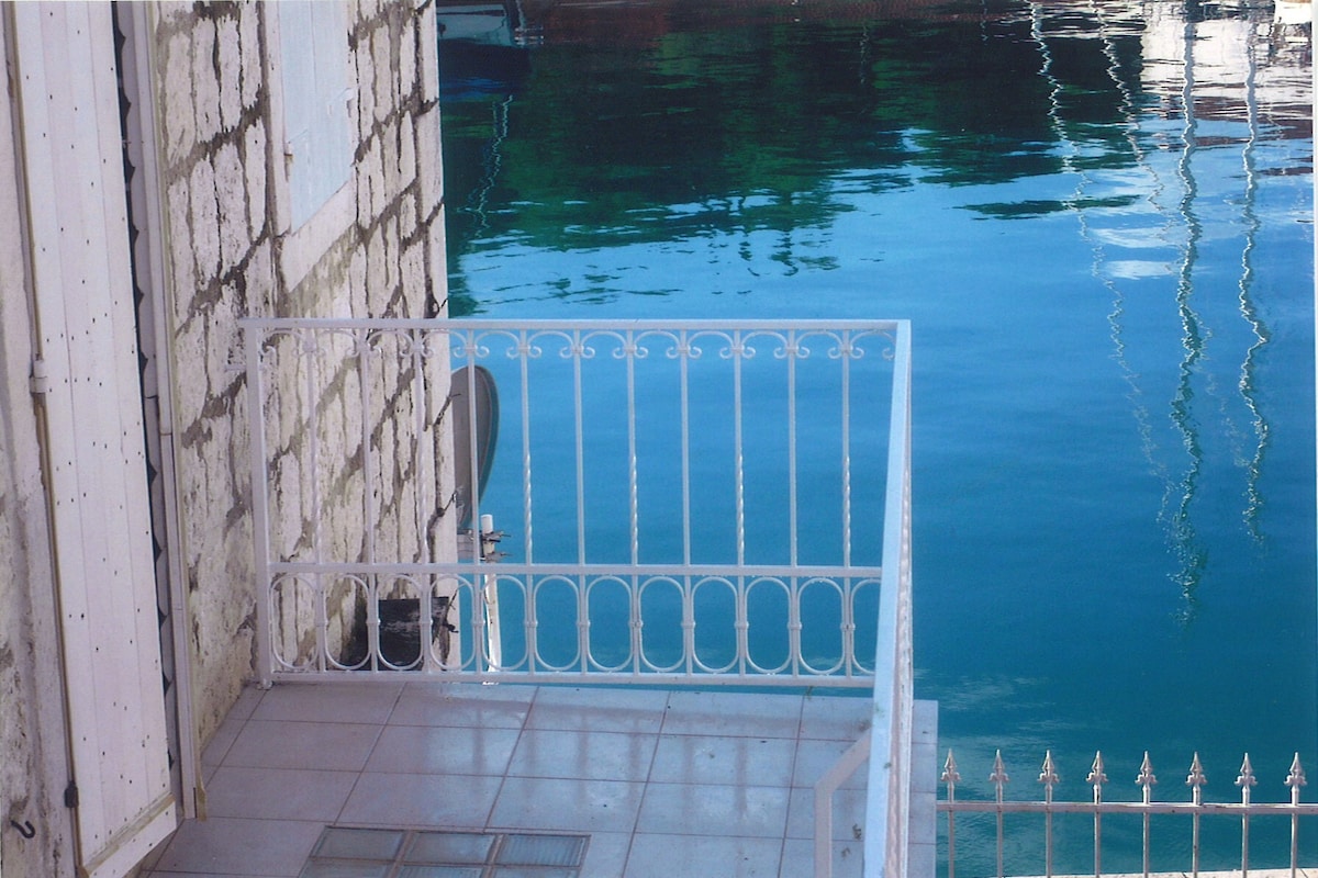 A white wrought-iron railing surrounds a small terrace overlooking clear blue waters. The reflections of the surrounding area can be seen in the water's surface, adding a serene element to the setting.
