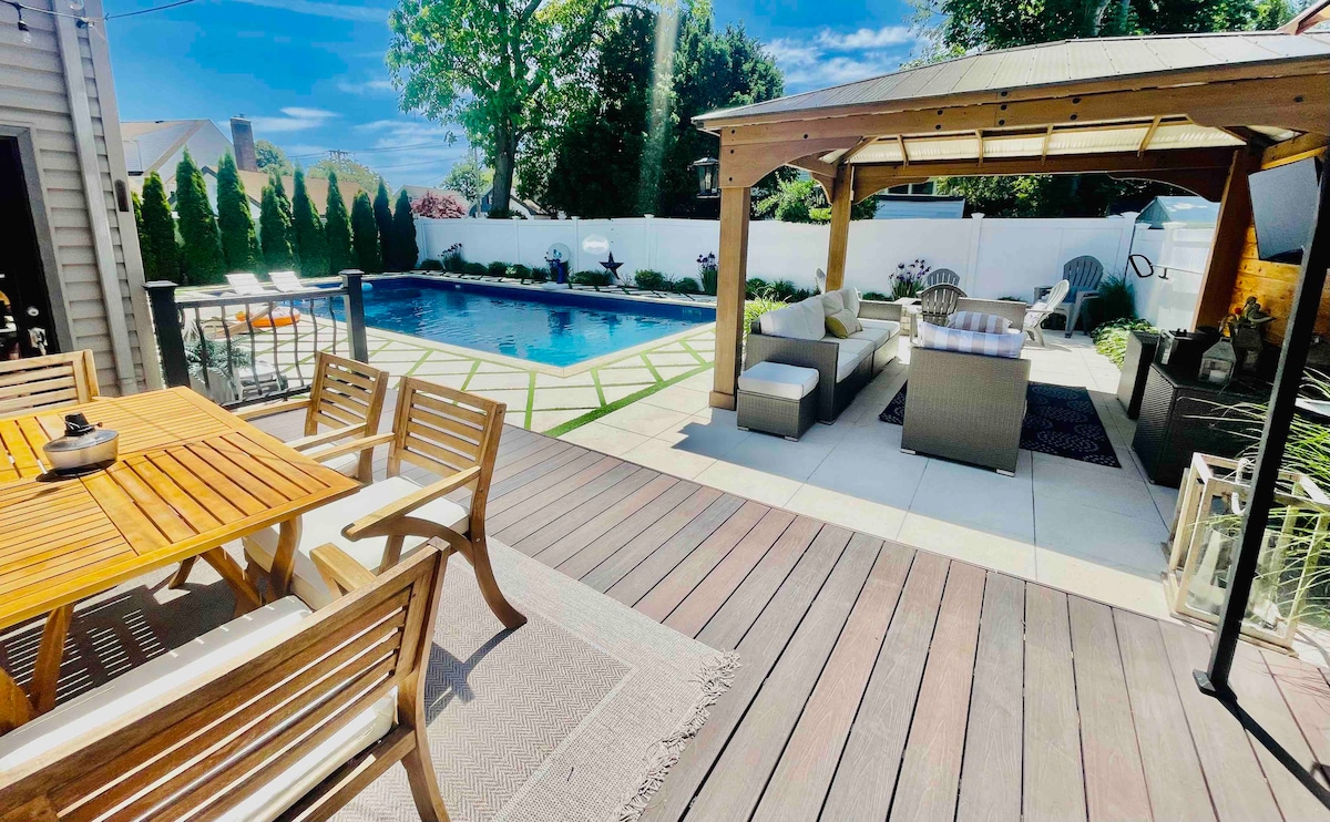 An outdoor space features a wooden dining table surrounded by six chairs, positioned on a deck. A lounge area with comfortable seating is seen beside an inground pool. The pool area is enclosed by a white fence and landscaped with greenery, contributing to a serene atmosphere.