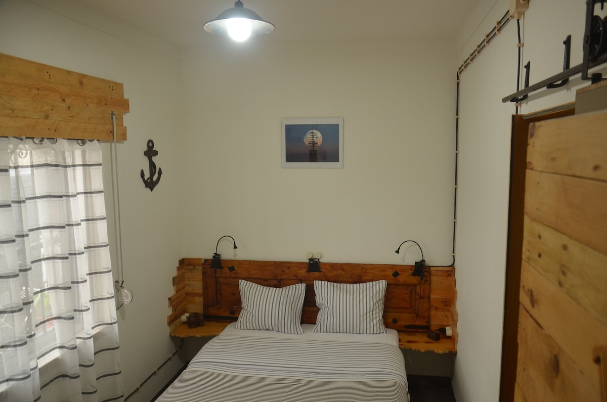 A cozy bedroom features a wooden headboard with matching bedside shelves. Soft lighting is provided by wall-mounted lamps on either side. A window is dressed with sheer curtains, allowing natural light to brighten the space, while a nautical-themed photo adorns the wall above the bed.