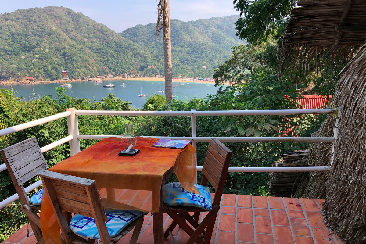A scenic view from a terrace showcases a small table with chairs, surrounded by vibrant greenery. The bay with boats is visible in the distance, framed by lush mountains. The warm tones of the tablecloth complement the natural surroundings.