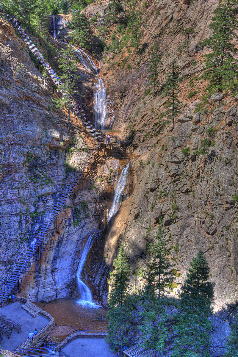 A cascading waterfall tumbles down rocky cliffs, surrounded by tall pine trees. Meandering paths and wooden stairs are visible, offering access to various viewpoints. The sunlight illuminates the rocky landscape, highlighting the natural beauty of the area.