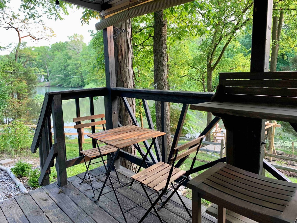 A wooden deck features a small table surrounded by two folding chairs. The view includes lush greenery and the calm surface of a lake, creating a serene outdoor setting that invites relaxation.