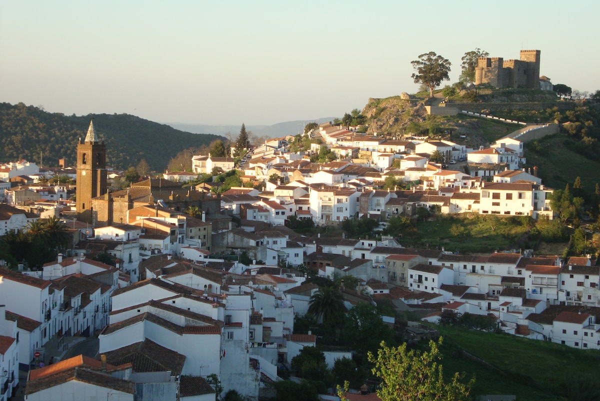 A panoramic view captures the quaint village nestled in the Sierra de Huelva, featuring white-washed buildings with terracotta roofs. In the background, a castle overlooks the landscape, surrounded by lush green hills and a clear sky, creating a serene countryside scene.