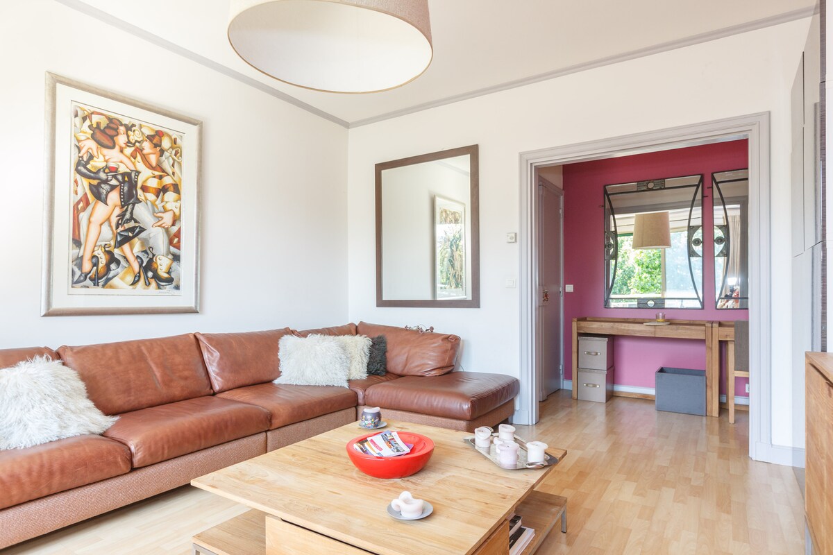 A comfortable living area features a brown leather sectional sofa, accented with white cushions. A wooden coffee table displays magazines and decorative items. The space is brightened by a large window and is complemented by a framed artwork and a large mirror on the wall.