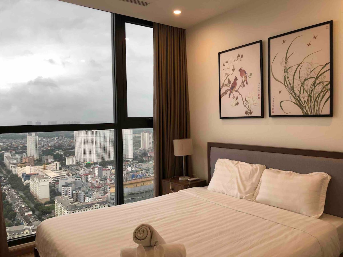 A modern bedroom is presented with large windows offering a city view. Two framed artworks depicting birds and plants adorn the wall above a spacious bed with white linens. A lamp is positioned beside the bed, complementing the neutral decor.