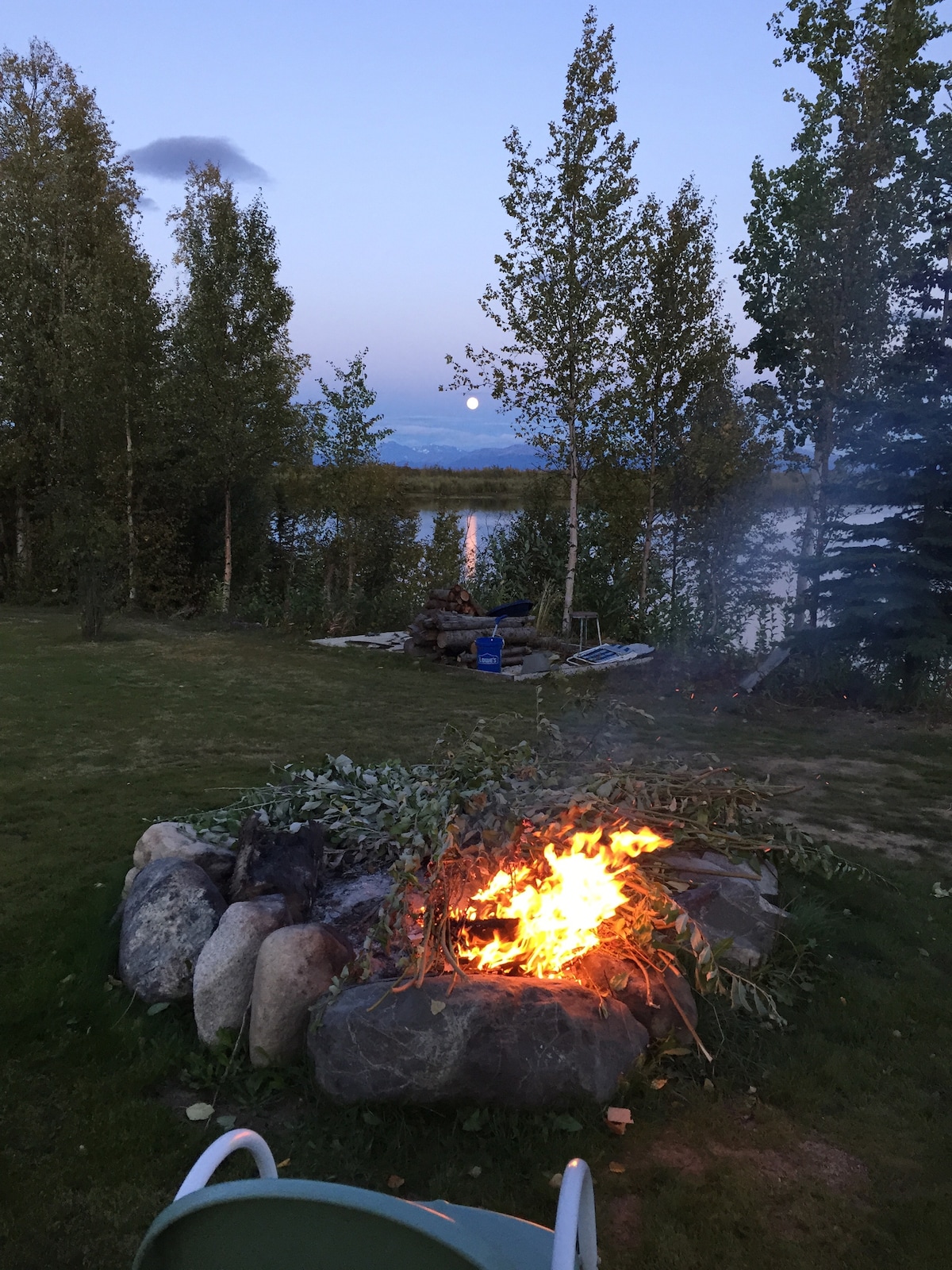 A fire pit made of large rocks is situated near Horseshoe Lake, surrounded by lush grass and trees. Flames flicker above a bed of leaves, emitting a gentle smoke. In the background, a serene lake reflects the evening sky, while a hint of the moon is visible.