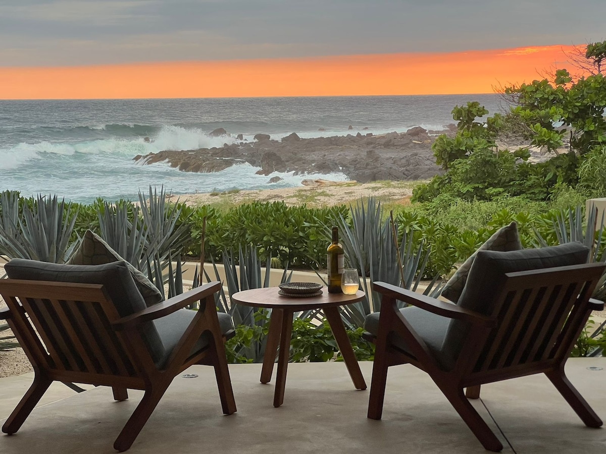 Two wooden lounge chairs are positioned on a patio, facing the ocean where waves crash against rocks. A small round table rests between the chairs, holding a bottle of wine. Lush greenery surrounds the area, while a warm sunset casts vibrant colors in the sky.