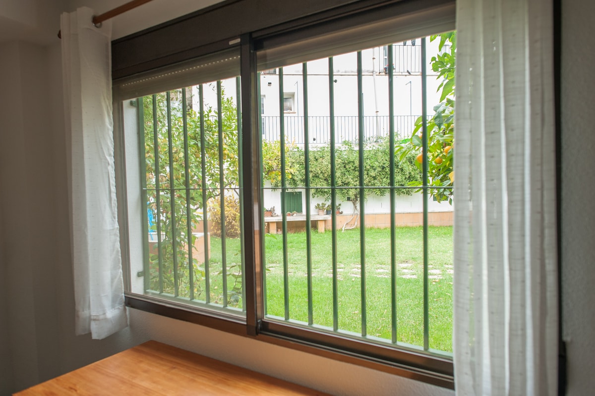 A window framed by light curtains offers a view of a serene garden. Lush greenery is visible outside, with well-maintained grass and scattered flower beds. Sunlight filters through the window, creating an airy atmosphere in the room.