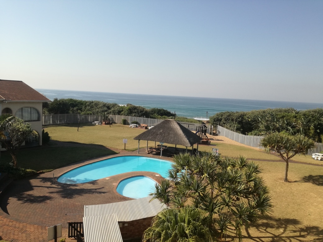 A tranquil outdoor area is highlighted by a clear blue swimming pool surrounded by lush green grass. Beach views extend in the background, with a thatched gazebo visible beside the pool. The ocean can be seen gently meeting the shoreline in the distance.