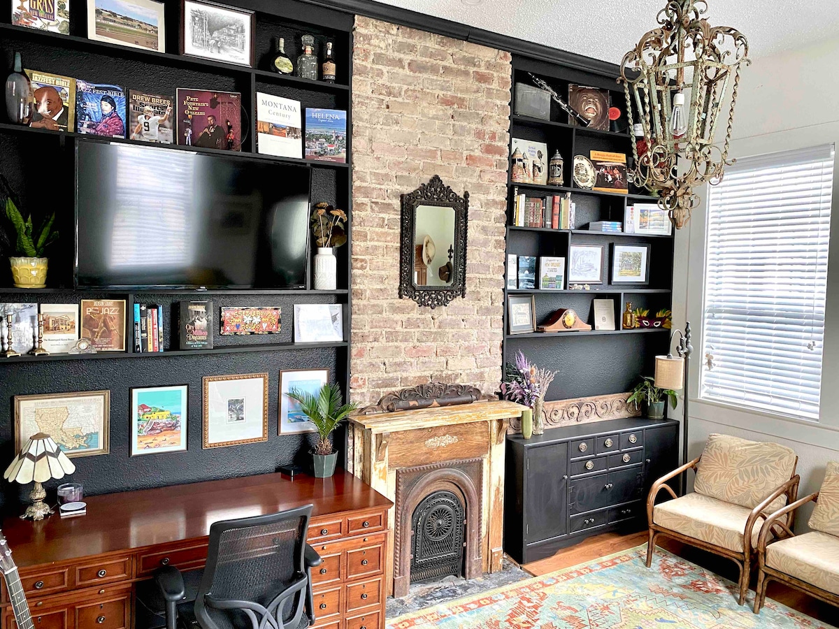 A cozy living area is highlighted by a brick feature wall, showcasing a stylish blend of books and art. A dark wooden desk and chair provide a workspace, while a unique fireplace mantel adds character. Light filters through nearby windows, complemented by a vintage chandelier.