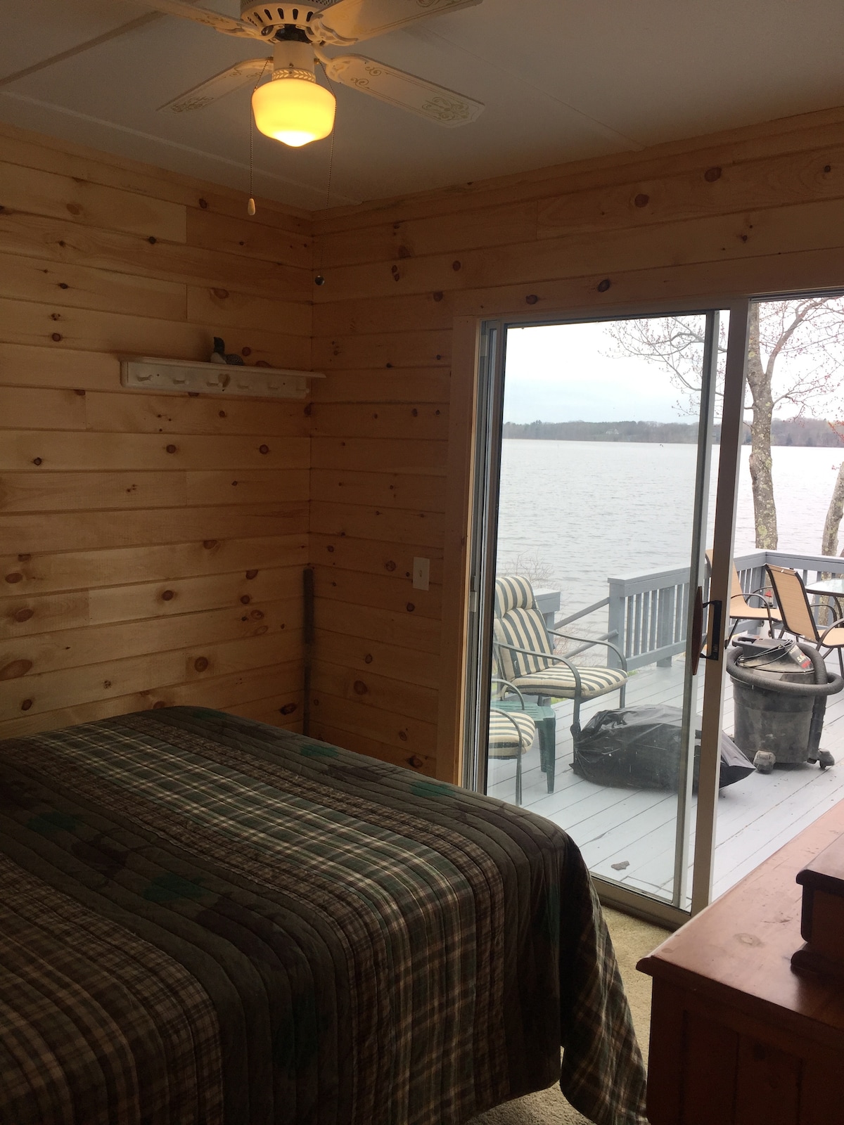 A wooden-paneled bedroom features a cozy bed with a plaid coverlet. Sliding glass doors open to a deck with seating, offering views of the water and nearby trees. Natural light fills the space, creating a serene atmosphere.