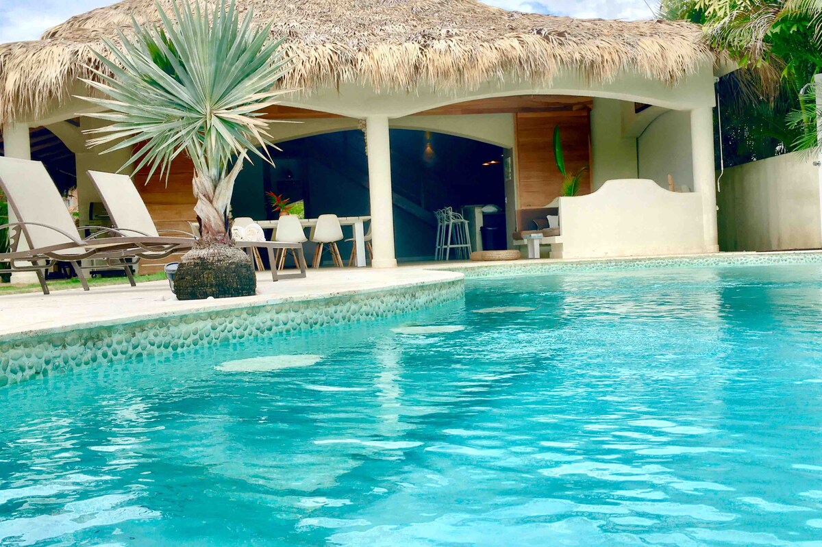 A private pool is framed by lush greenery, with four lounge chairs positioned nearby. The pool's clear blue water reflects the thatched roof of the outdoor lounge area, which includes a dining space and a view of the interior living area.