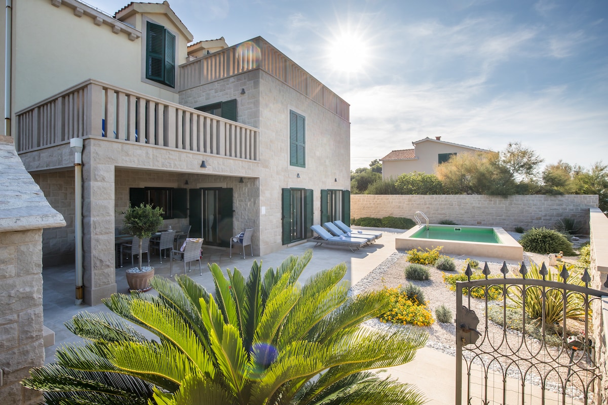 The villa's exterior showcases a spacious patio area with lounge chairs positioned near a small pool. Lush greenery and colorful landscaping enhance the inviting space. The building features large glass doors and windows, allowing natural light to fill the interior.