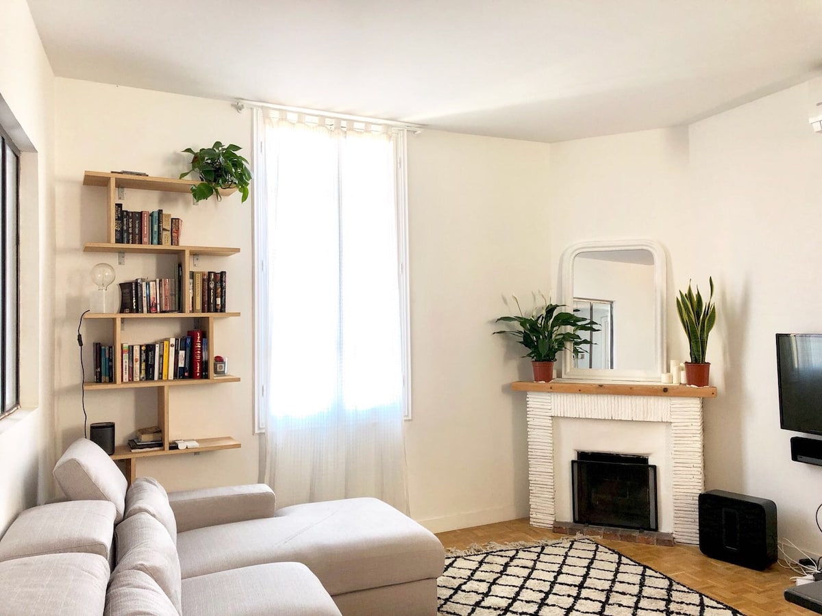 A spacious living area is presented, featuring a light-colored sectional sofa and a textured area rug. A bookshelf filled with books is positioned against one wall, while the fireplace, framed by a mirror, is adorned with potted green plants. Natural light filters through the large window.