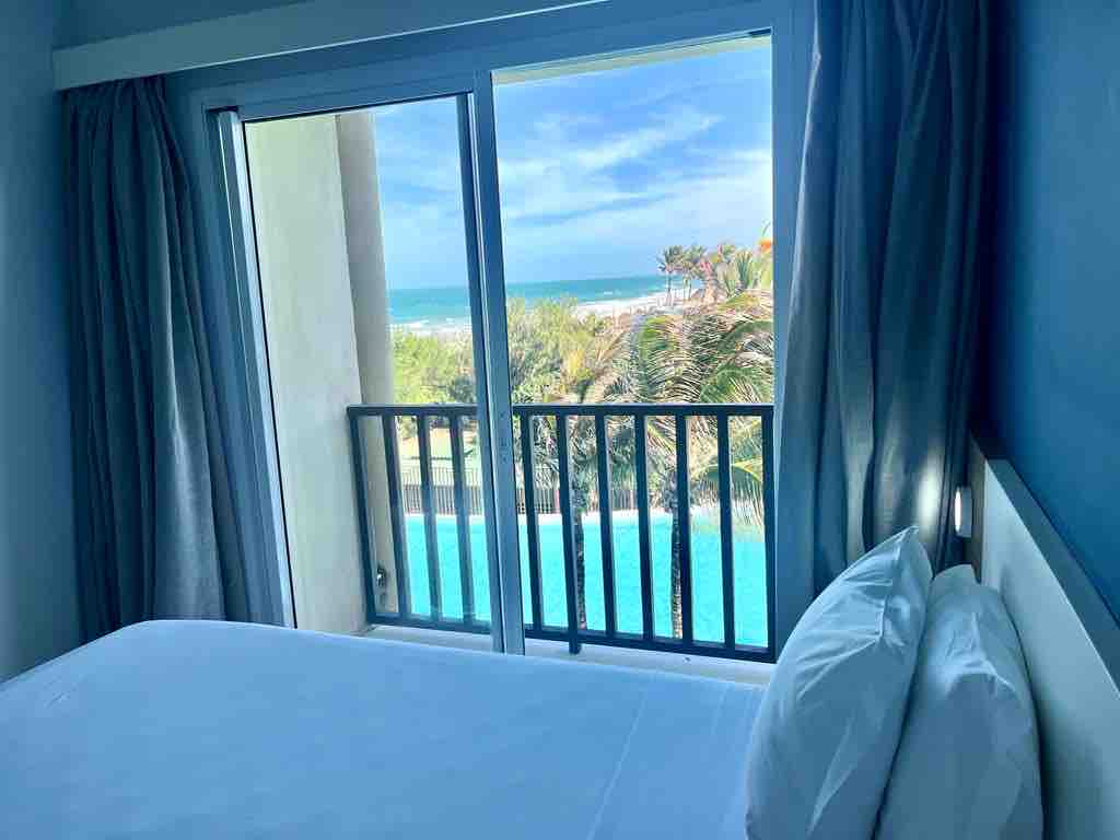 A serene bedroom is depicted with a spacious bed featuring white linens. A balcony door provides a view of the ocean and palm trees, with bright natural light filling the room. Soft grey curtains frame the doorway, enhancing the calm atmosphere.