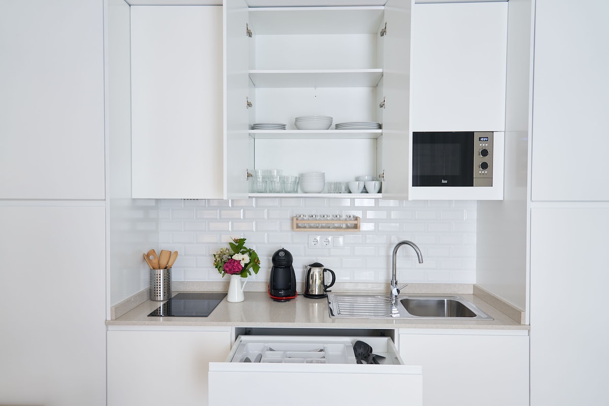 A modern kitchen features white cabinetry with open shelves displaying dishware. A stainless steel sink and an electric kettle are visible, alongside a coffee maker and a compact microwave. A bouquet of flowers adds a touch of color to the countertop.