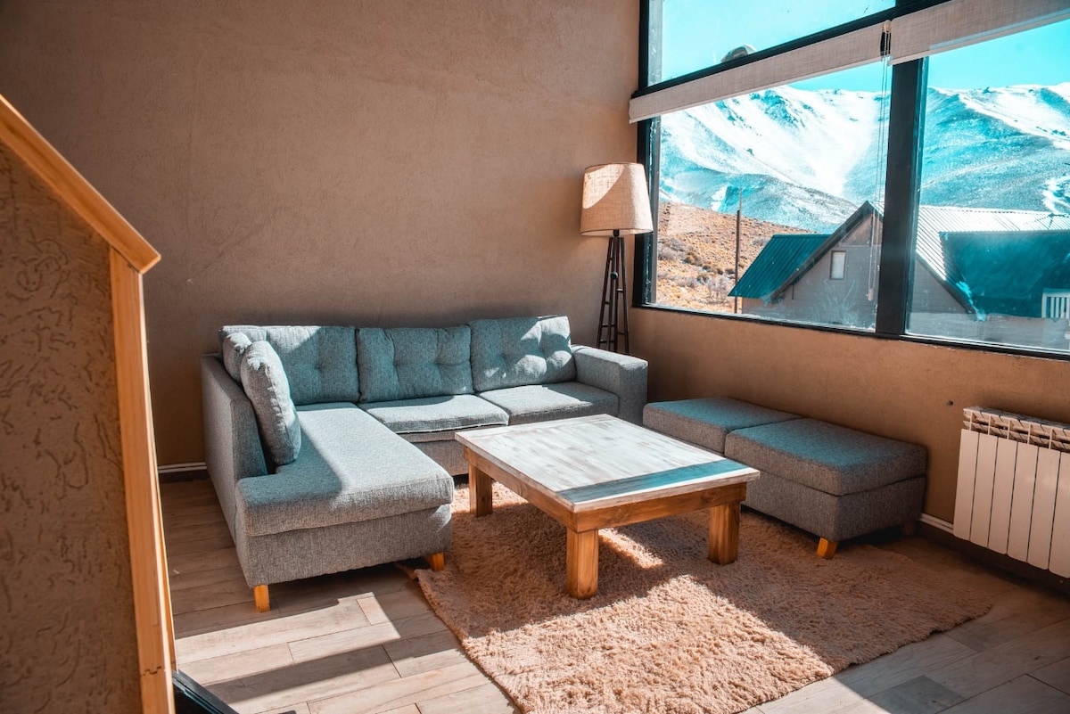 A spacious living area features a sectional sofa complemented by a matching ottoman. A wooden coffee table occupies the center, while large windows frame scenic views of the mountains, allowing for ample natural light to fill the room, with a cozy rug underfoot.