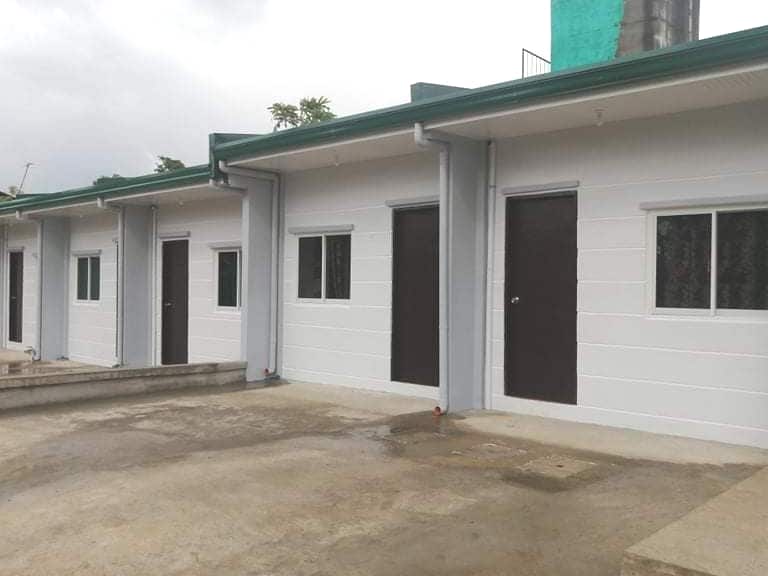 A row of simple, modern units is seen with neutral-colored facades. Each unit features a dark door and a small window beside it. The concrete pathway leads to a clean, open area in front, surrounded by greenery.