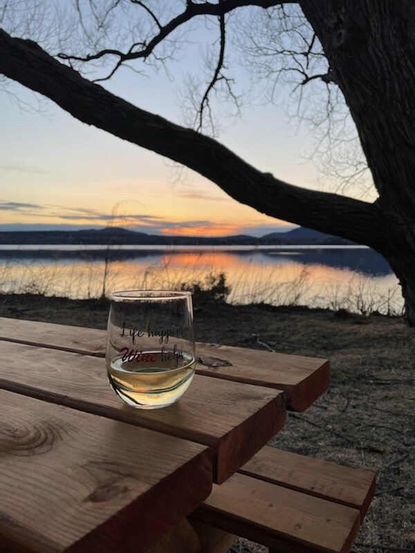 A wooden picnic table with a clear glass of wine is placed in the foreground. In the background, a sunset paints the sky with warm tones, reflecting on the calm surface of the water, surrounded by trees.