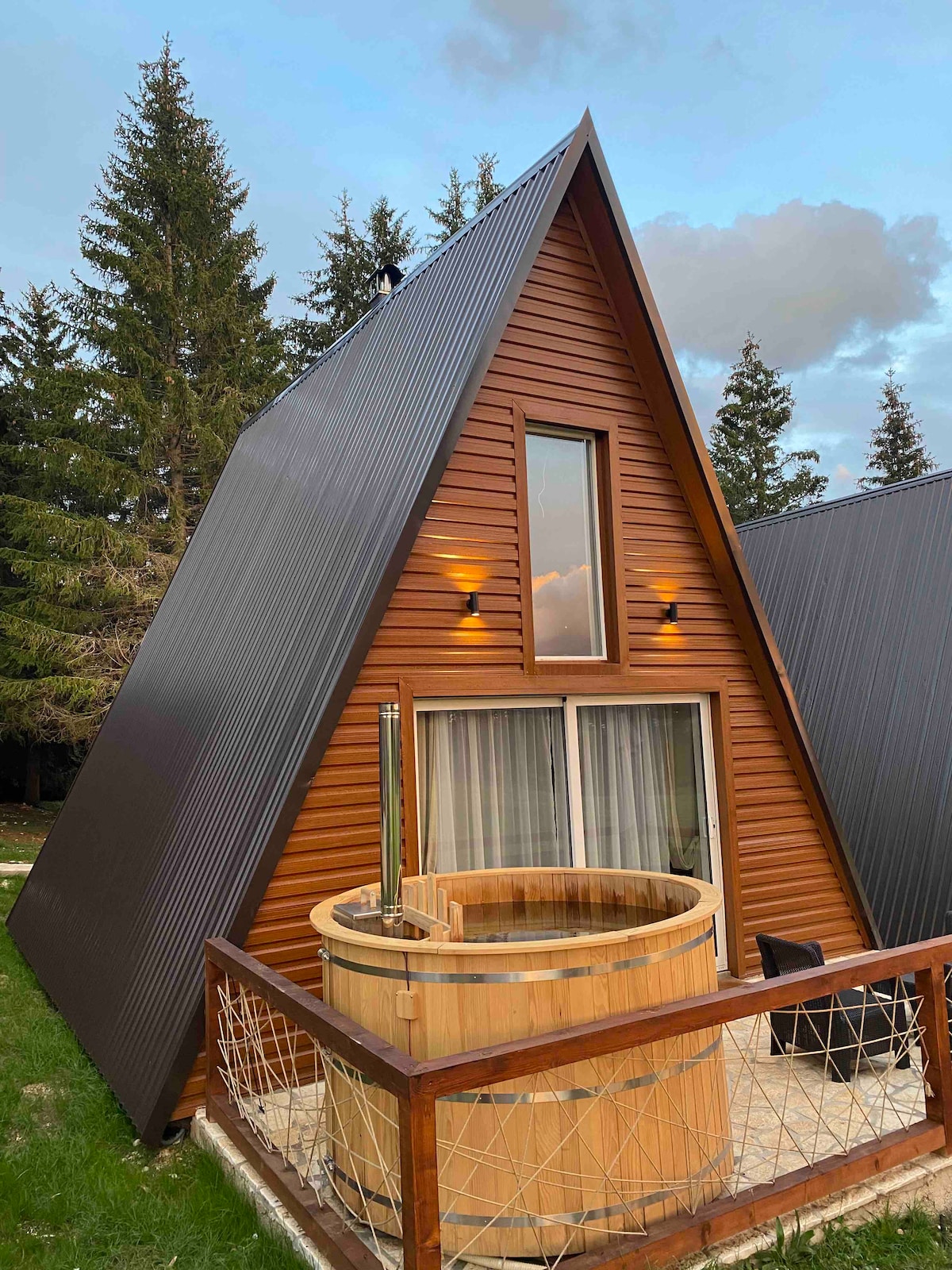 An A-frame cabin constructed with wooden panels is highlighted by large windows. A wooden hot tub is positioned on a private terrace, complemented by natural landscaping featuring tall evergreen trees in the background.