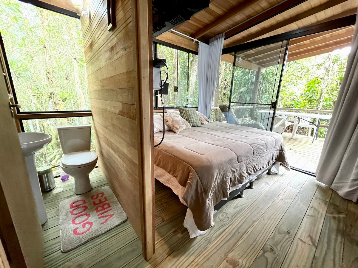 A section of a wooden cabin features a spacious bed adorned with neutral linens. Natural light filters through large glass doors, connecting indoor and outdoor spaces. A private bathroom area is visible, alongside a rug with the text 'GOOD VIBES'. Lush greenery is seen outside.