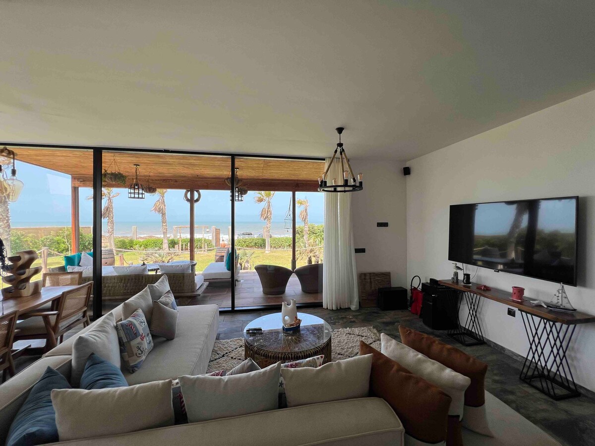 A spacious living area is presented, featuring a comfortable sectional sofa and a round coffee table atop a neutral rug. Large glass doors offer views of the outdoor space and ocean, while natural light brightens the room. A wall-mounted television and a decorative chandelier are visible.