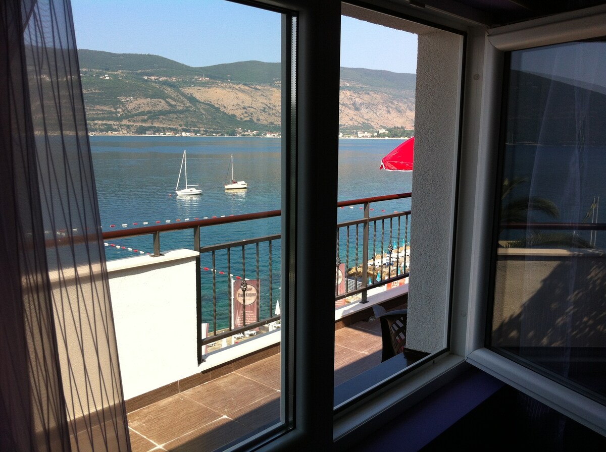 A scenic view of the sea is framed by large windows, showcasing calm waters and distant mountains. Sailboats are anchored in the foreground, while a red umbrella adds a pop of color to the balcony area outside. Soft curtains allow natural light to filter into the room.