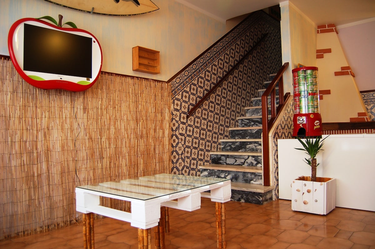 A reception area features a tiled floor and a staircase leading to the upper level. A glass-topped table with wooden legs sits in the center. Decorative elements include a wall-mounted television and a colorful vending machine. A potted plant adds a touch of greenery.