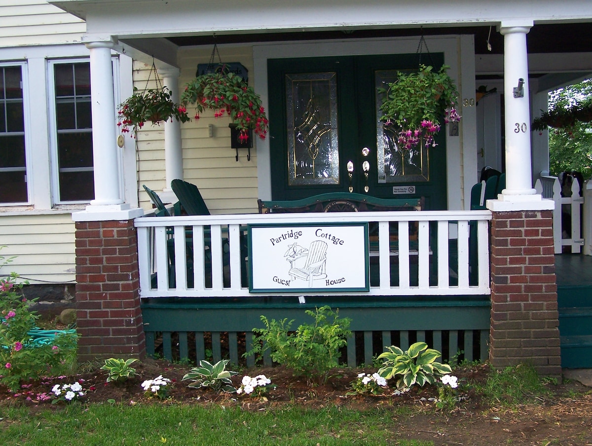 The entrance to Partridge Cottage is framed by hanging flower baskets and a welcoming front porch. Comfortable Adirondack chairs are positioned on the deck, inviting relaxation. A decorative sign with an illustration of a chair complements the charming exterior, which is finished with a white and green color scheme.