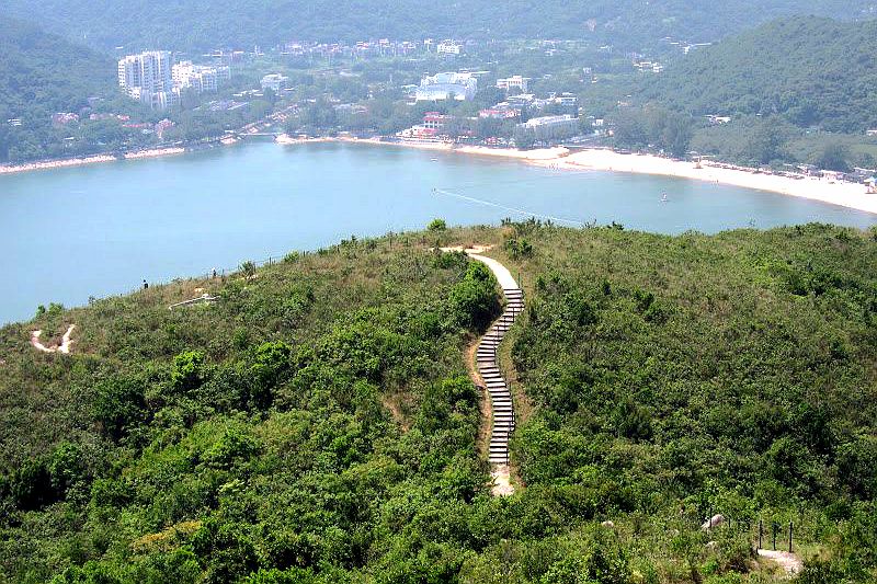 A scenic view of a coastal area showcases a winding trail leading down through lush greenery towards the shoreline. The serene bay is visible in the background, bordered by soft sandy beaches and gently rolling hills that create a peaceful natural landscape.