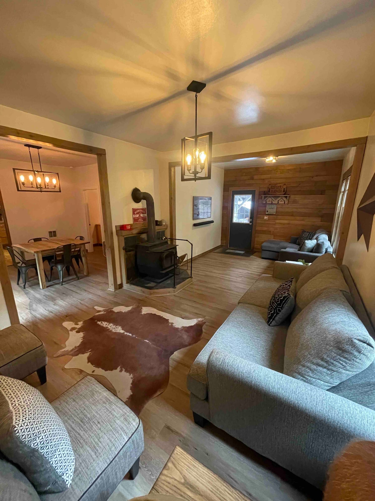 A spacious living area features comfortable seating arranged around a decorative rug. A wood stove adds warmth, while natural light filters through a nearby window. An open doorway leads to a dining space, enhancing the layout of the cabin.