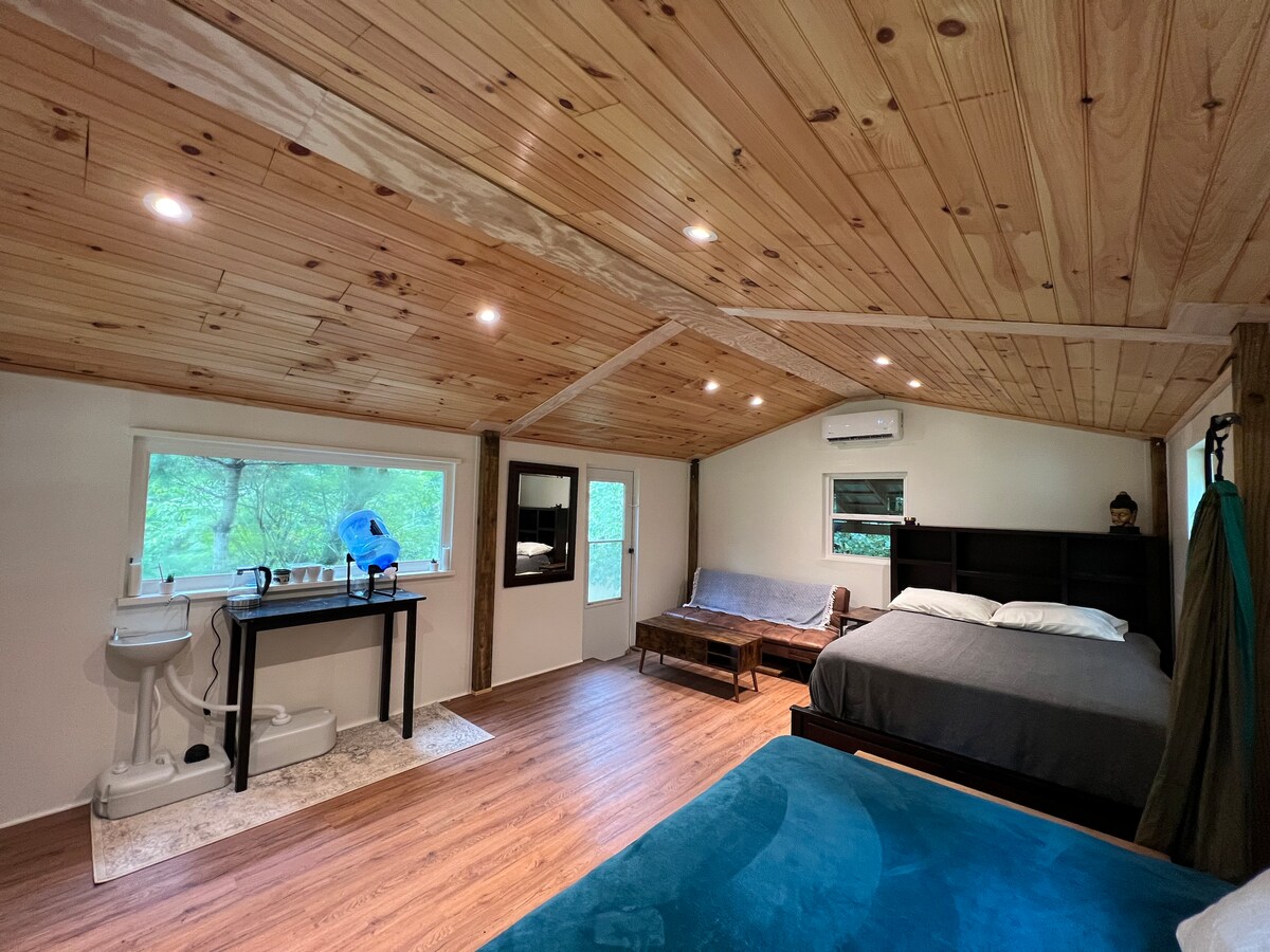 The open living space features a spacious queen bed and a full-sized bed, both draped in simple linens. Natural light pours through the windows, illuminating the wood-paneled ceiling. A kitchenette with basic amenities is present, along with a comfortable seating area designed for relaxation.