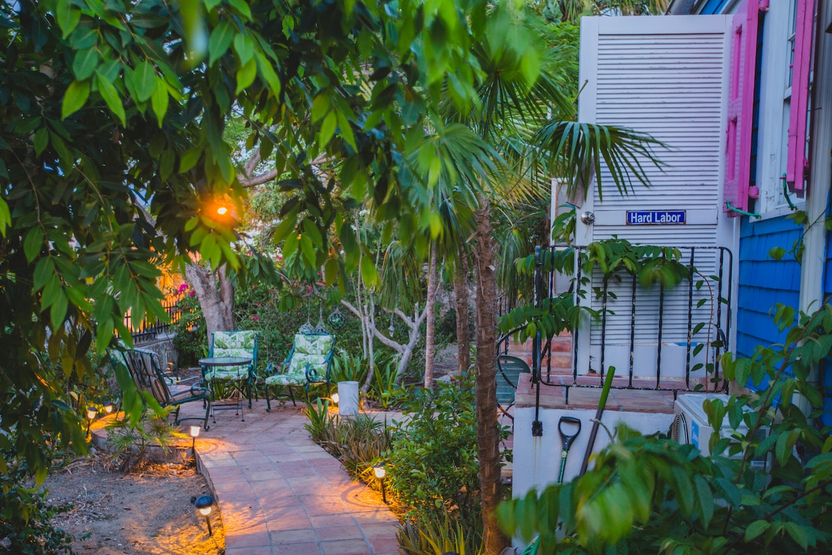 A pathway is surrounded by lush greenery and tropical plants, leading to a welcoming porch adorned with vibrant pink shutters. Soft outdoor lighting creates a serene ambiance, while patio furniture invites relaxation amidst the tranquil garden setting.