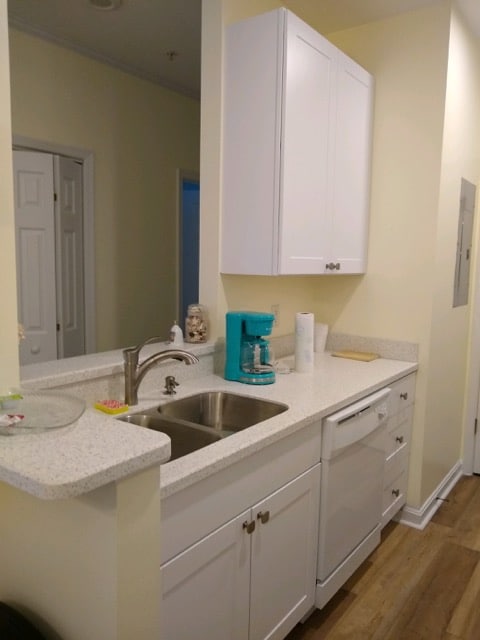 A neatly organized kitchen area is visible, featuring white cabinetry and a clean countertop. A stainless steel sink and dishwasher are integrated into the setup. A vibrant blue coffee maker adds a pop of color, while the light walls contribute to a bright atmosphere.