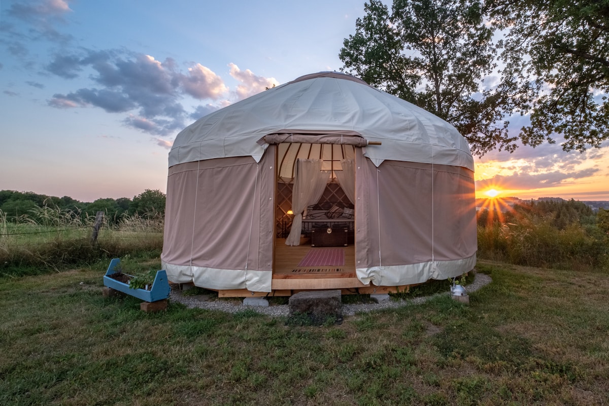 A stylish yurt is highlighted by the setting sun, creating a serene atmosphere. The entrance, framed with soft fabric, opens to reveal a cozy interior. Surrounding green hills and a grassy area provide a tranquil setting. Natural light enhances the yurt's inviting appearance.