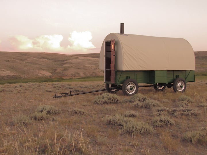 Heward's Fully Restored Sheep Wagon, a green old-fashioned wagon with tan canvas roof and sides with woodstove pipe on a prairie.