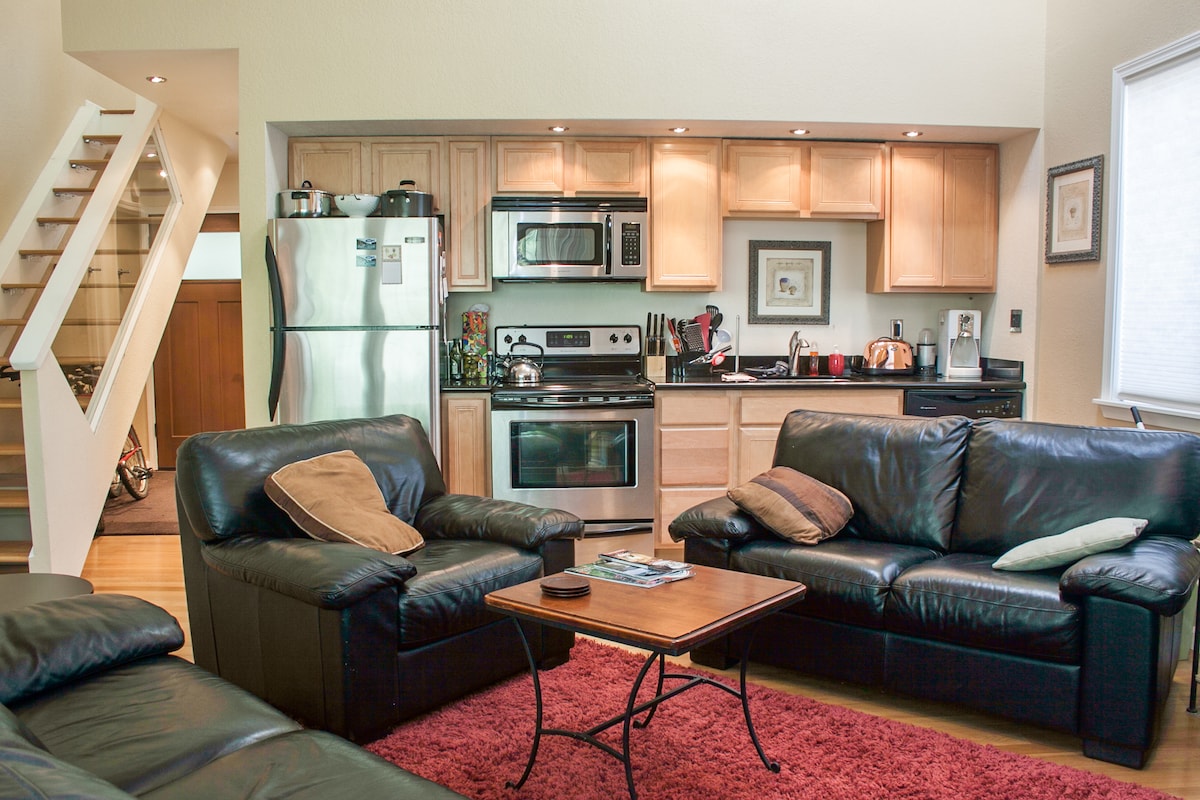A spacious living area features dark leather sofas paired with light-colored cushions. A wooden coffee table is centered among the seating. The bright kitchen is equipped with stainless steel appliances, including a refrigerator and microwave, with wooden cabinetry above and a staircase visible in the background.