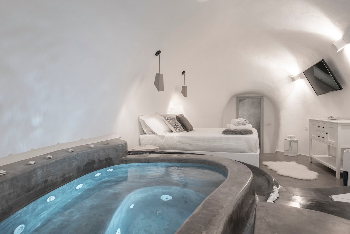 A spacious bedroom features a large bed with soft linens beside a rounded jacuzzi. The arched ceiling is complemented by minimalist light fixtures, and a flat-screen TV is mounted on the wall, providing a modern touch to the serene interior.