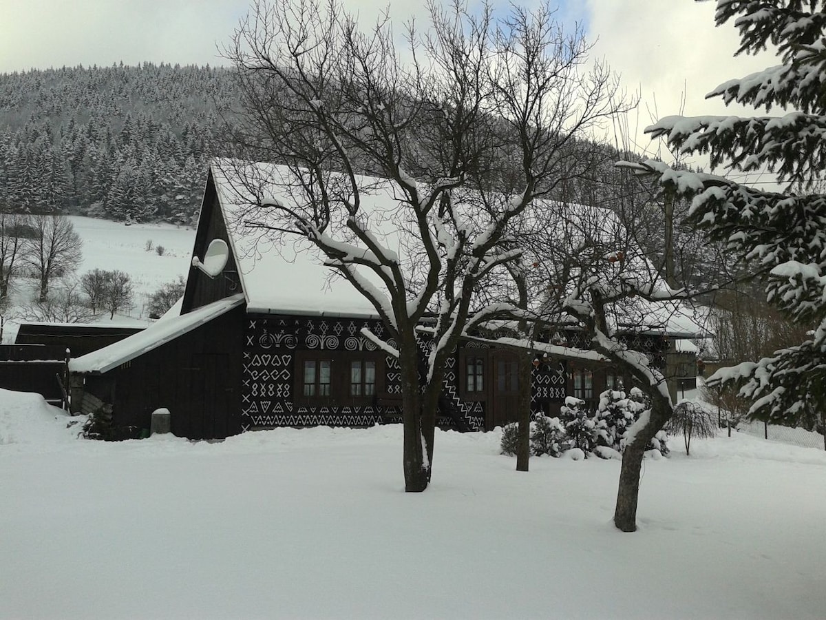 A traditional wooden cottage, adorned with intricate black and white patterns, rests amidst a snowy landscape. Nearby trees are dusted with snow, and a gentle slope of white accents the backdrop. The serene setting reflects a peaceful winter atmosphere.