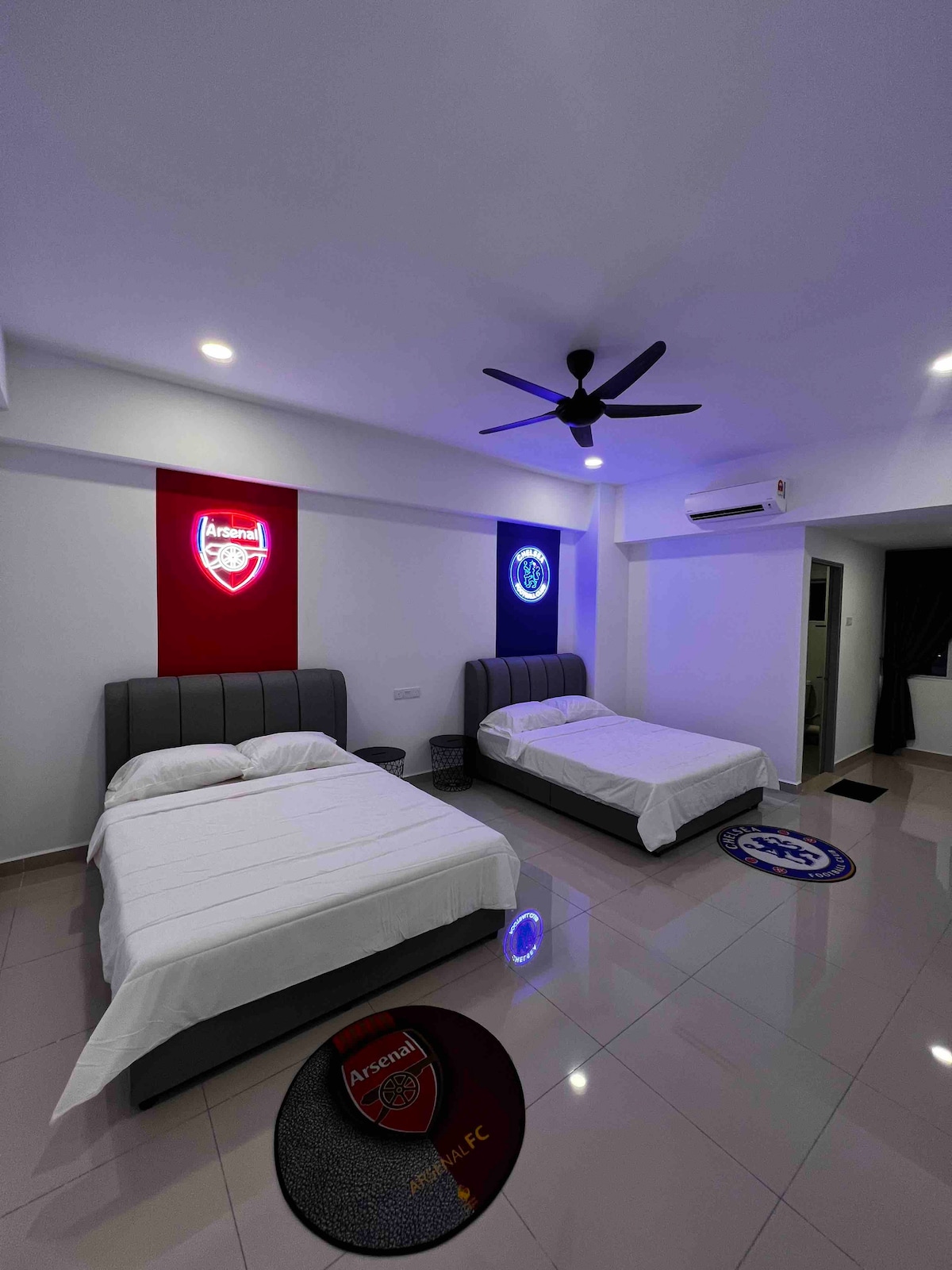 The room features two spacious beds, each dressed in white linens. Above the beds, illuminated wall art displays the Arsenal and Manchester City logos. A ceiling fan and sleek tile flooring create a modern ambiance, while a circular rug adds color to the space.