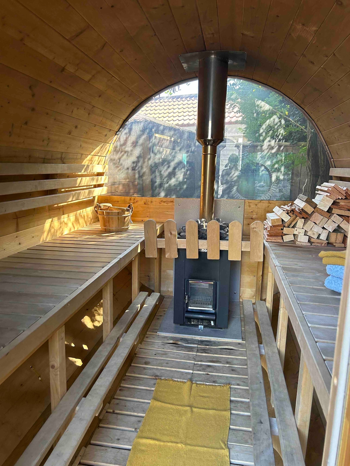 A wooden sauna interior features seating arranged on both sides, complemented by wooden shelves and a central wood-burning stove. The soft glow of natural light enters through the curved walls, illuminating the warm wooden tones. A small pile of firewood rests beside the stove, adding to the rustic ambiance.