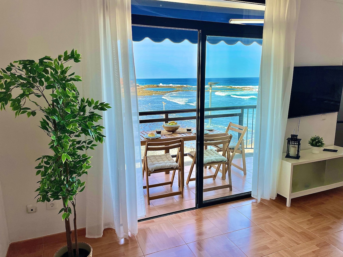 Large glass doors open to a furnished balcony overlooking the ocean, where a small table and two chairs are positioned. Light white curtains frame the view, allowing natural light to filter into the spacious living area. The sea and coastal scenery are visible beyond.