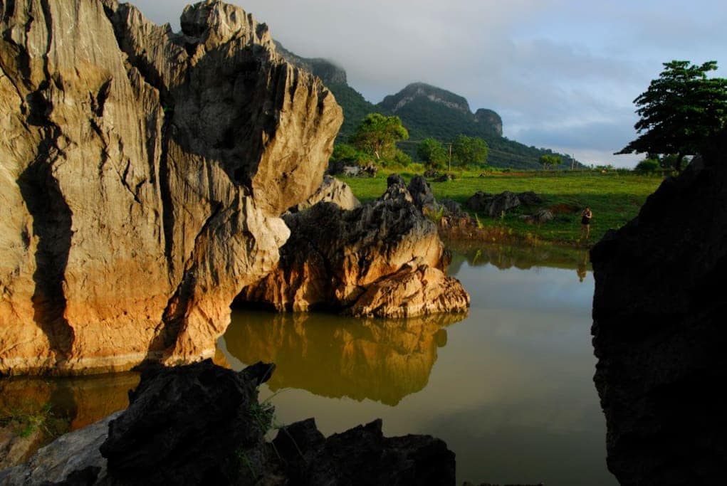 A serene landscape is depicted featuring large, rugged rocks surrounding a calm body of water. Lush greenery is visible in the background, with gentle slopes leading to distant mountains partially covered by clouds. The scene conveys a peaceful natural environment.