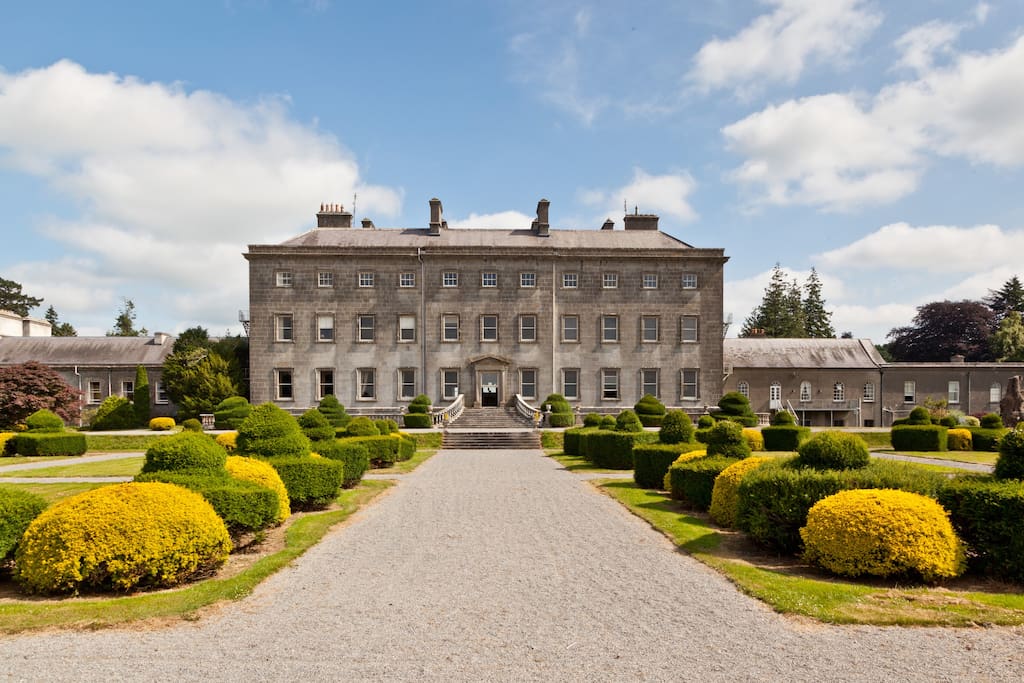 a large stone building with a walkway and bushes with Powerscourt Estate in the background