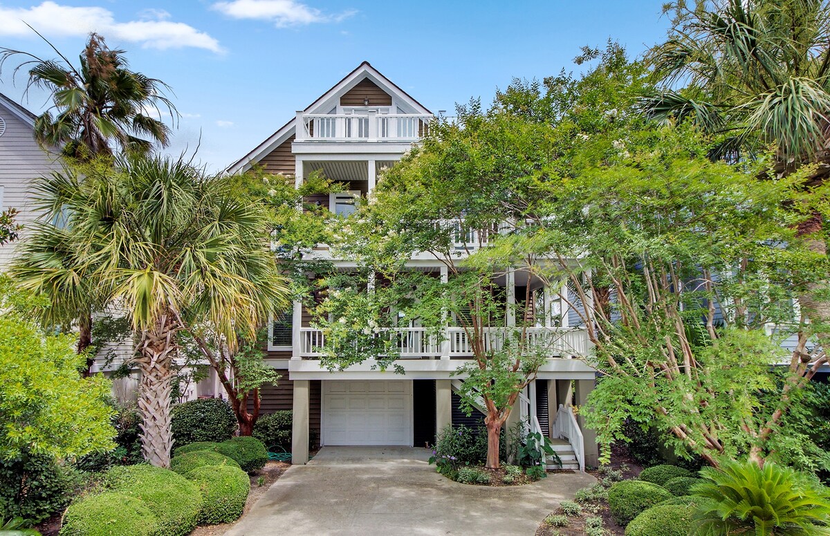 The three-story home is surrounded by lush greenery and palm trees. The exterior features a broad porch and multiple balconies, inviting relaxed outdoor living. A driveway is visible leading to a garage, while well-maintained landscaping enhances the overall appearance of the property.