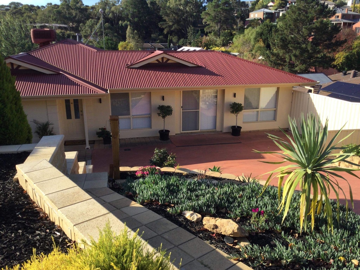 A charming exterior view of a single-story home with a red roof, surrounded by neatly landscaped gardens. Pathways are lined with flowering plants and shrubs, leading up to the main entrance which is framed by two small trees. The setting offers a serene atmosphere.